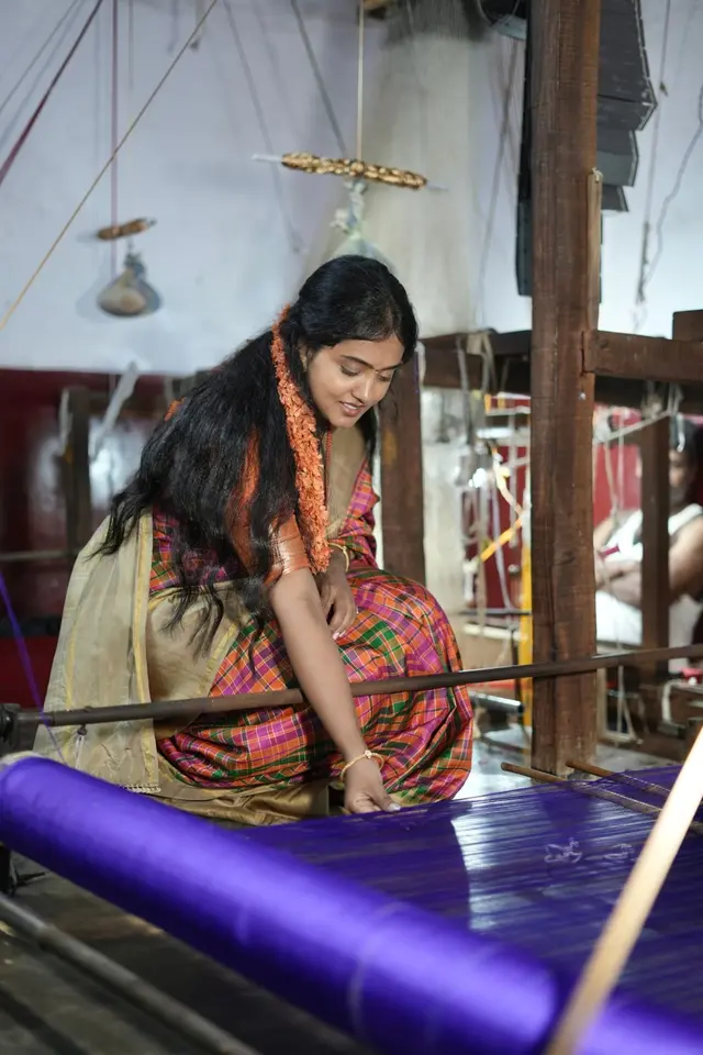 Gowri reaching for the purple silk warp on a loom, a weaver at work behind her