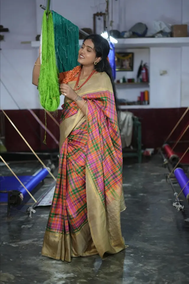 Gowri, founder of Aradhya Silks, holding hanks of green silk yarn in a weaving unit, in an orange, pink and green checked silk saree